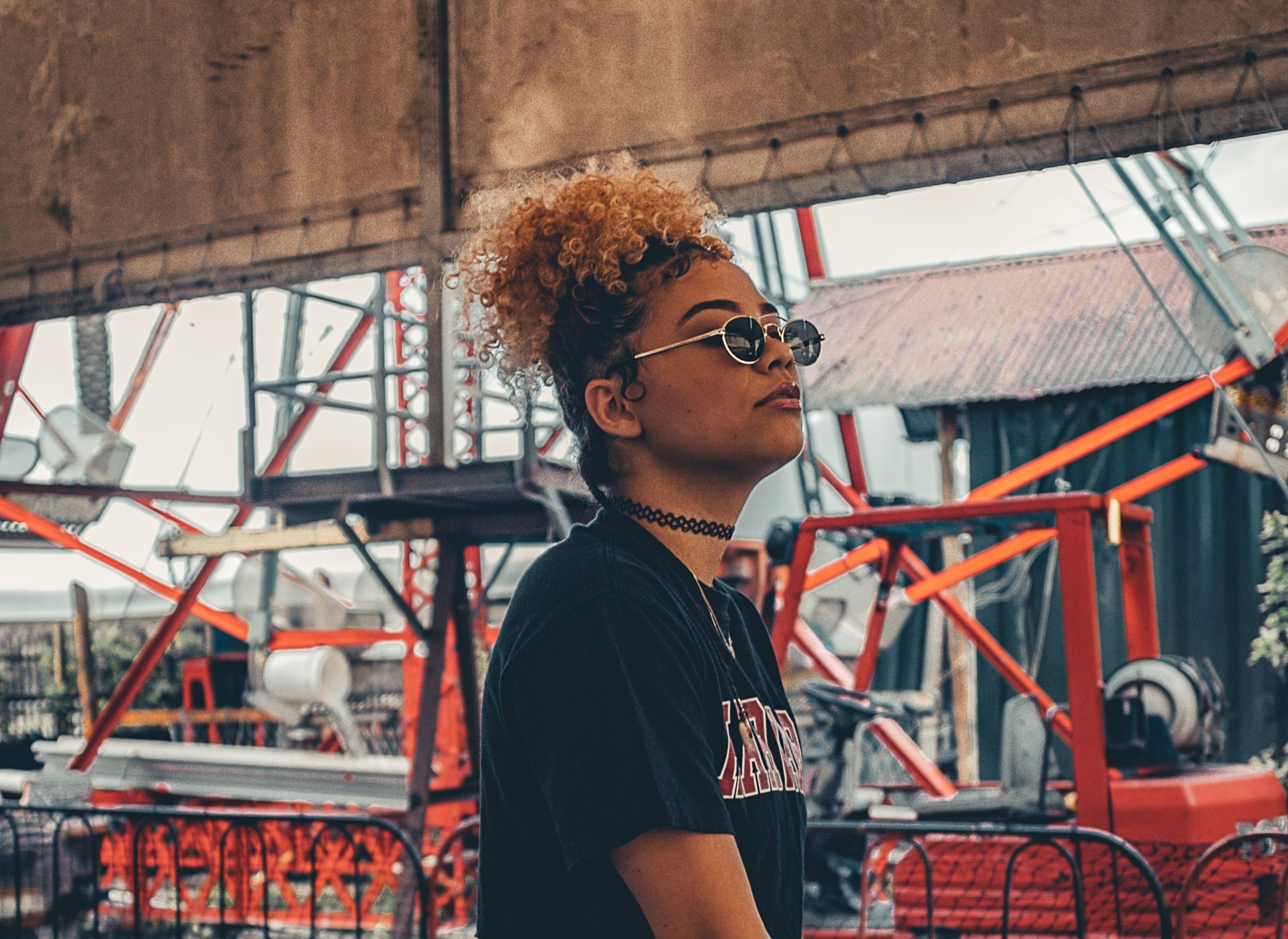  Mulher negra com cabelo encaracolado loiro e óculos de sol, perfil, com fundo industrial