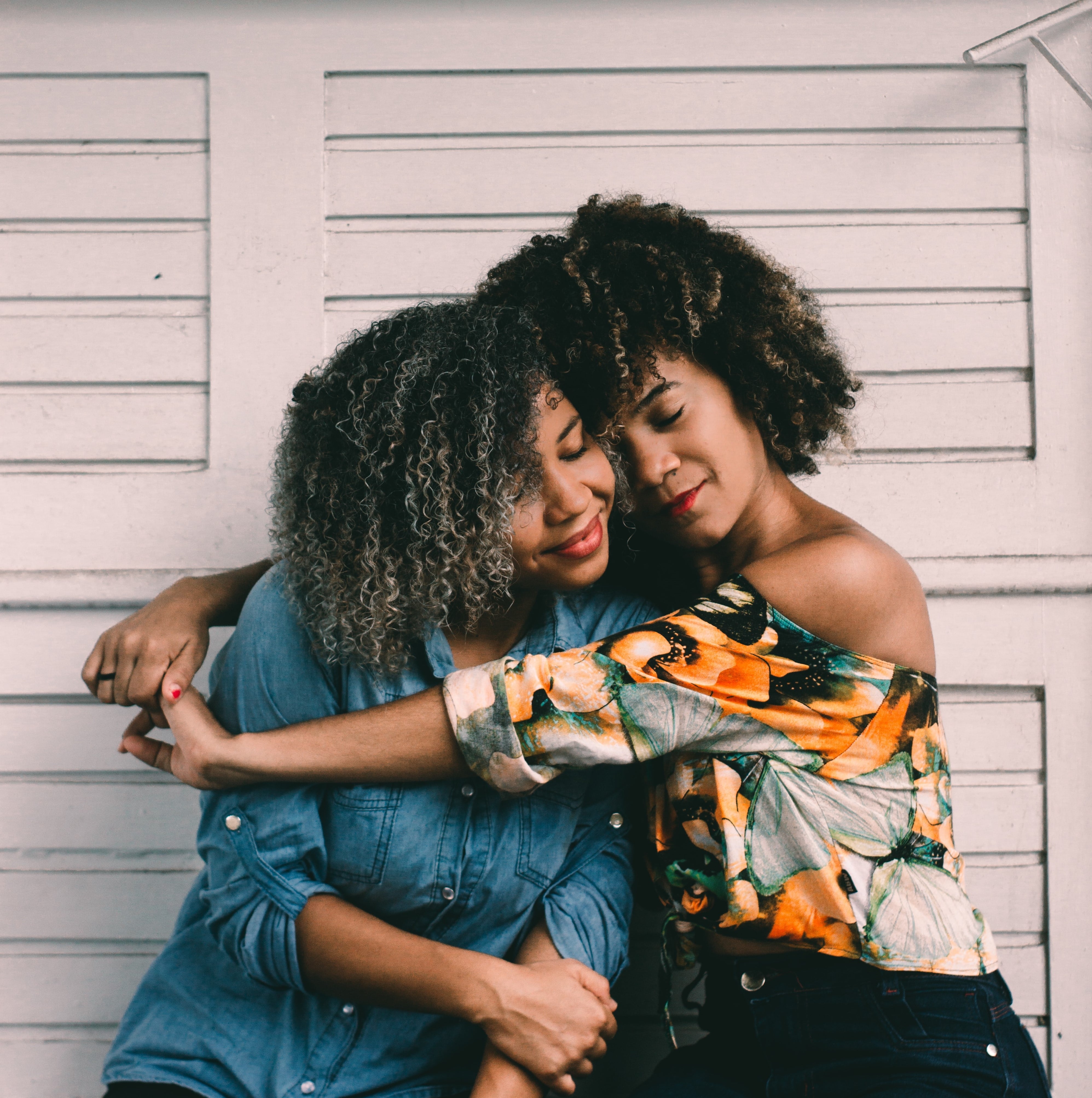  Mulheres abraçadas, olhos fechados; uma com jaqueta, a outra com blusa estampada, fundo de concreto desenhado