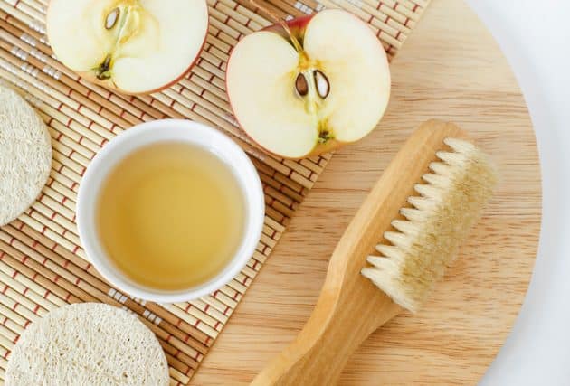 A imagem mostra uma maça partida ao meio, um recipiente com vinagre de maçã e uma escova de cabelo