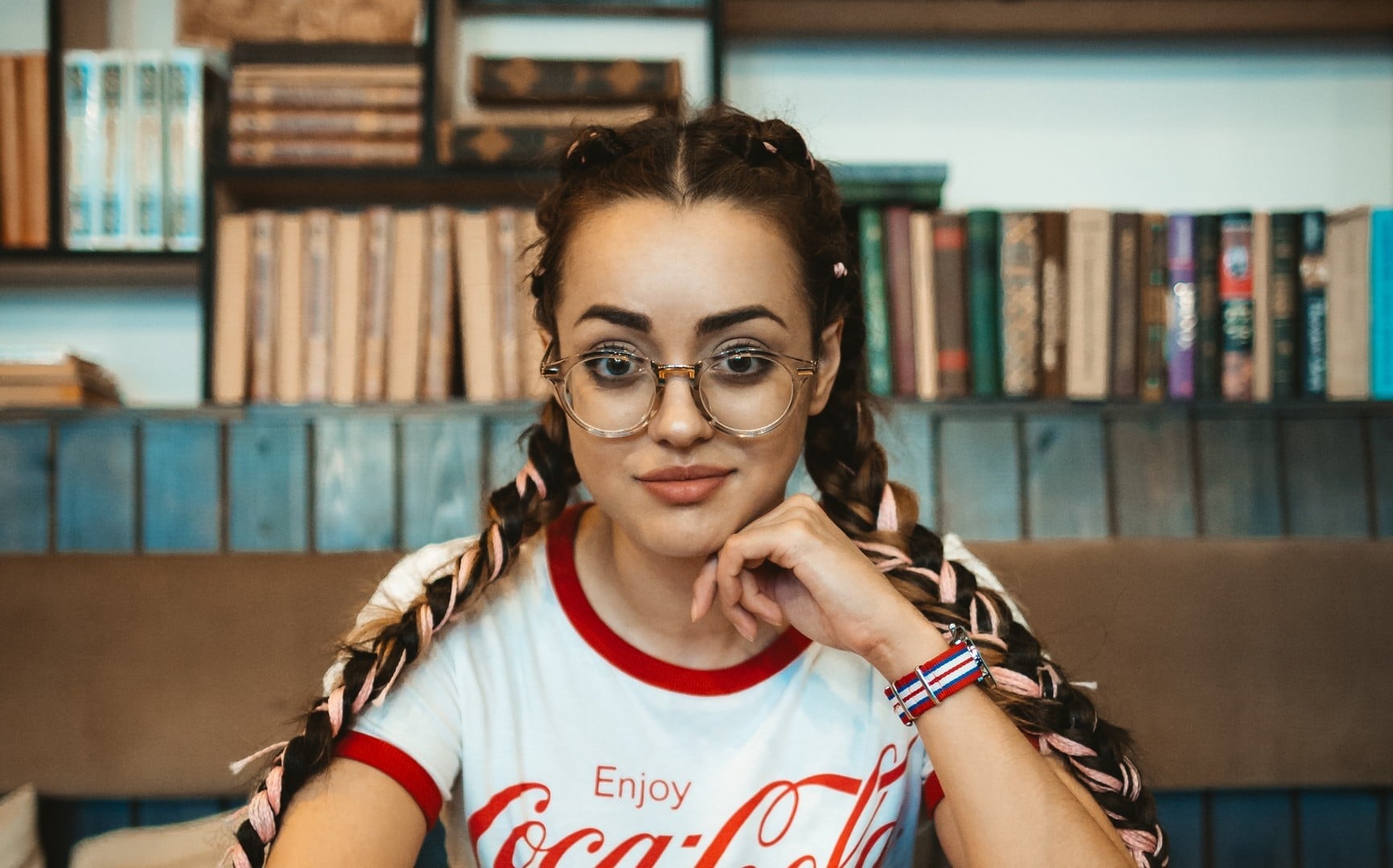  Mulher branca com óculos redondos e trança, mão no queixo, usando blusa branca da Coca-Cola com detalhes vermelhos