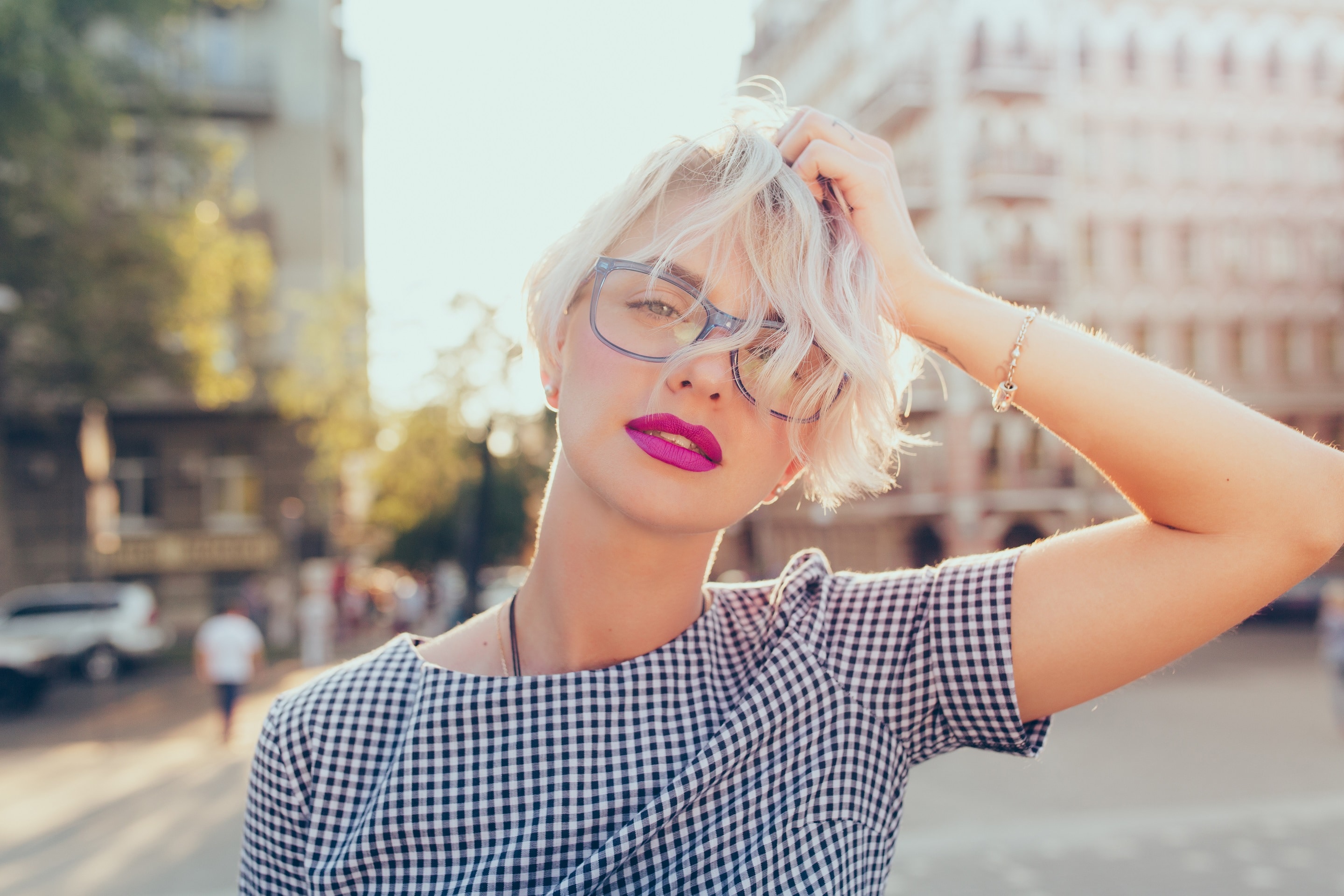  Mulher com cabelo curto loiro, óculos e batom rosa em ambiente urbano