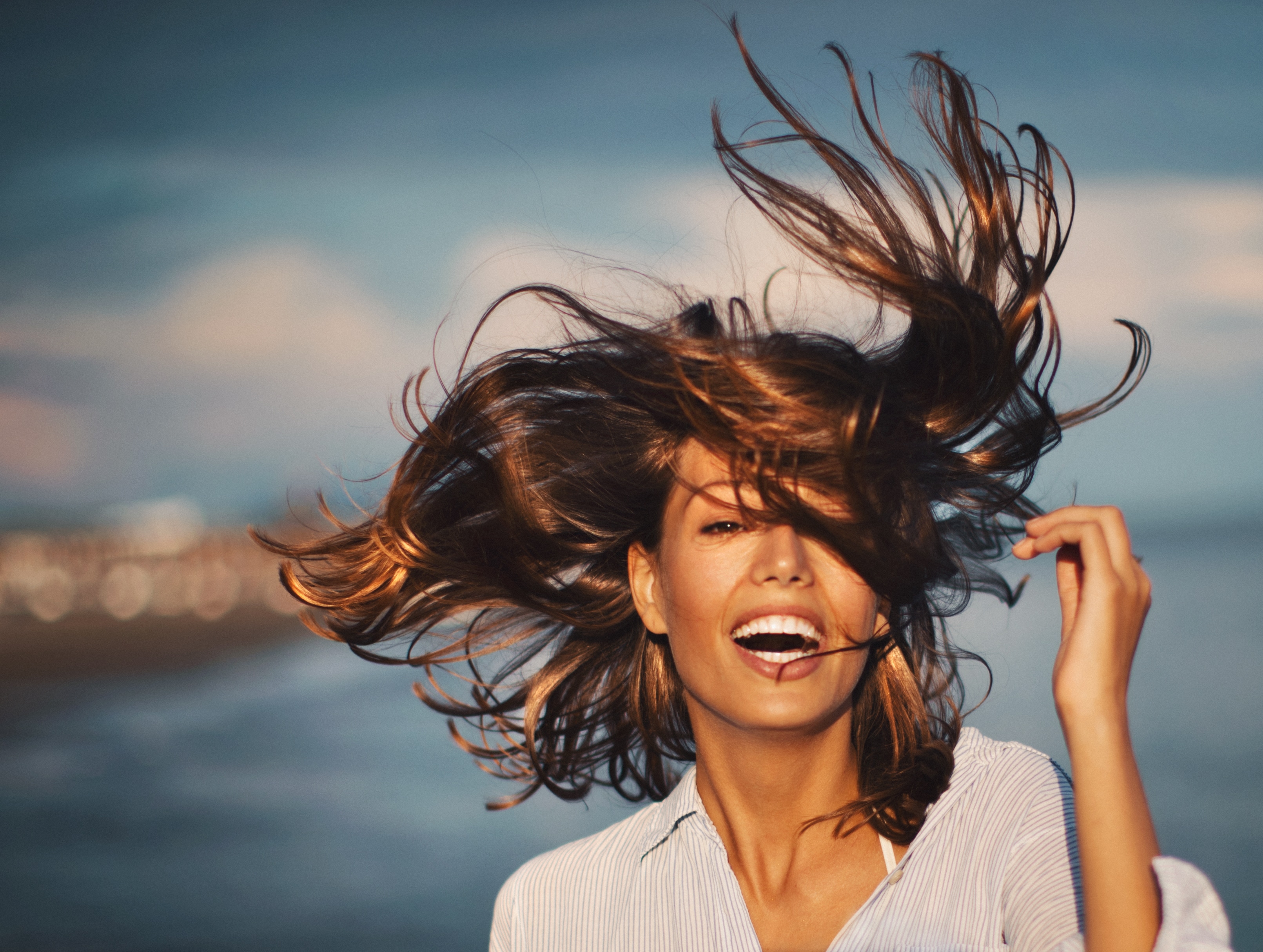  Mulher sorrindo com cabelo castanho ao vento e camisa listrada à beira-mar