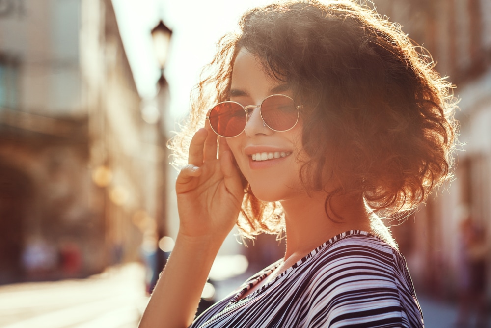  Mulher com óculos redondos e cabelo cacheado sorrindo ao ar livre