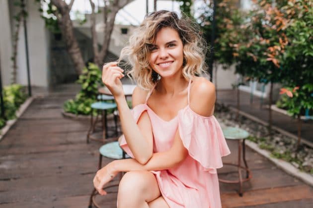  Mulher sorrindo com cabelo loiro cacheado e vestido rosa ombro a ombro sentada ao ar livre