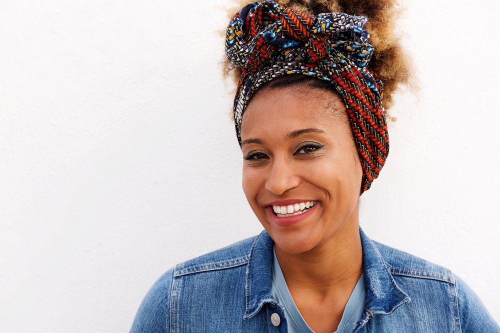  Mulher negra sorridente, com lenço colorido na cabeça, cabelos cacheados e jaqueta