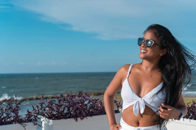 Mulher negra de cabelo liso, preto e longo, com progressiva. Ela está na praia, de costas para o mar e usa um óculos escuro