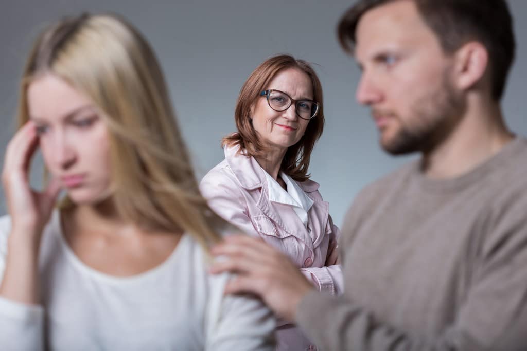  Um casal, na qual a mulher está brava, o homem tenta acalma-la e a sogra, atrás, da risada