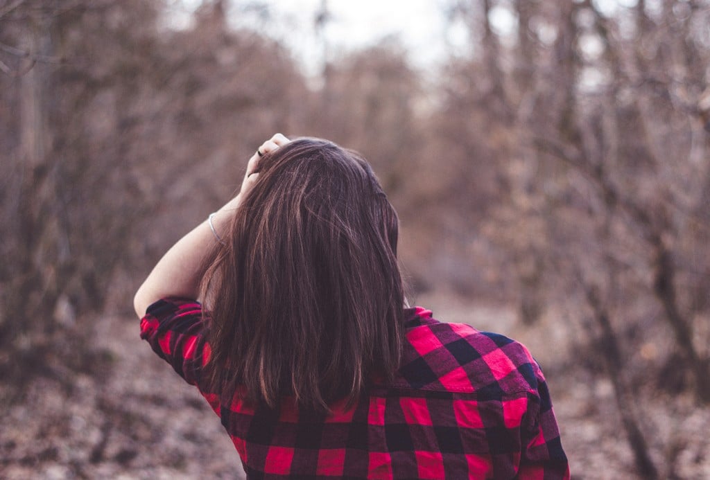  Mulher de camisa xadrez vermelha e preta, de costas, tocando o cabelo em ambiente natural com árvores secas ao fundo