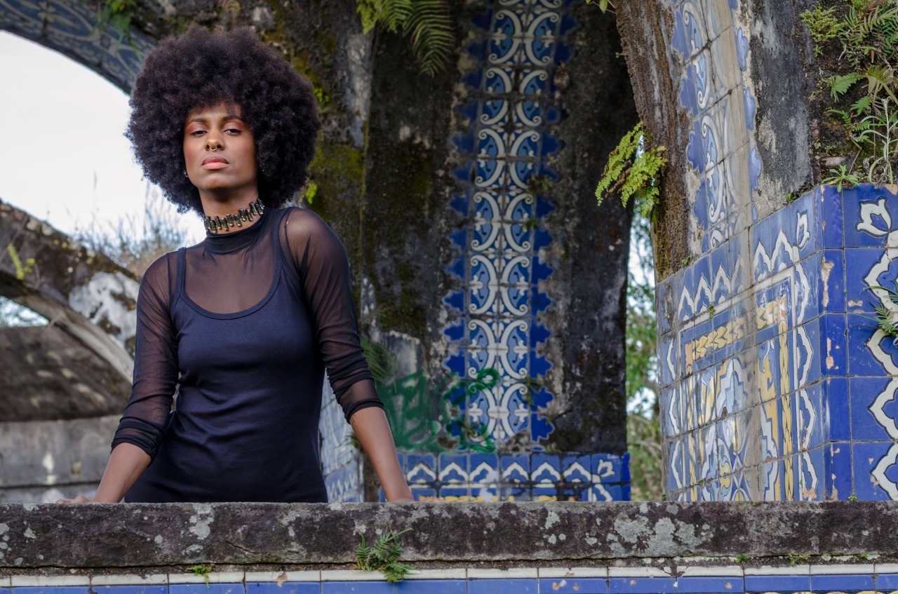  Mulher com cabelo afro volumoso, blusa preta, diante de azulejos azuis tradicionais