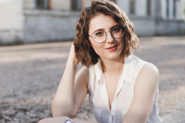 Mulher jovem com óculos redondos e cabelo curto ondulado em blusa branca, ao ar livre