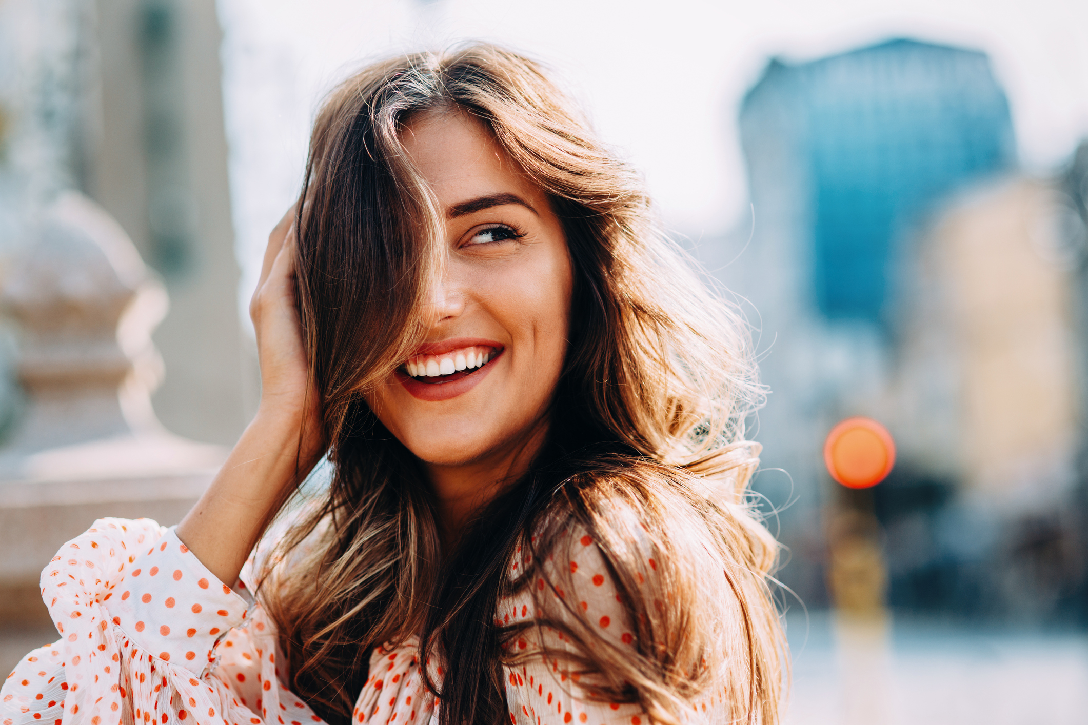  Mulher sorrindo, mão no rosto, cabelo tampando um dos olhos, cabelo ondulado claro e blusa estampada de bolinhas