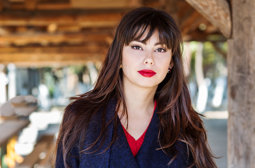  Mulher com cabelo longo, batom vermelho, blusão azul com blusa vermelha por baixo, e fundo de madeira