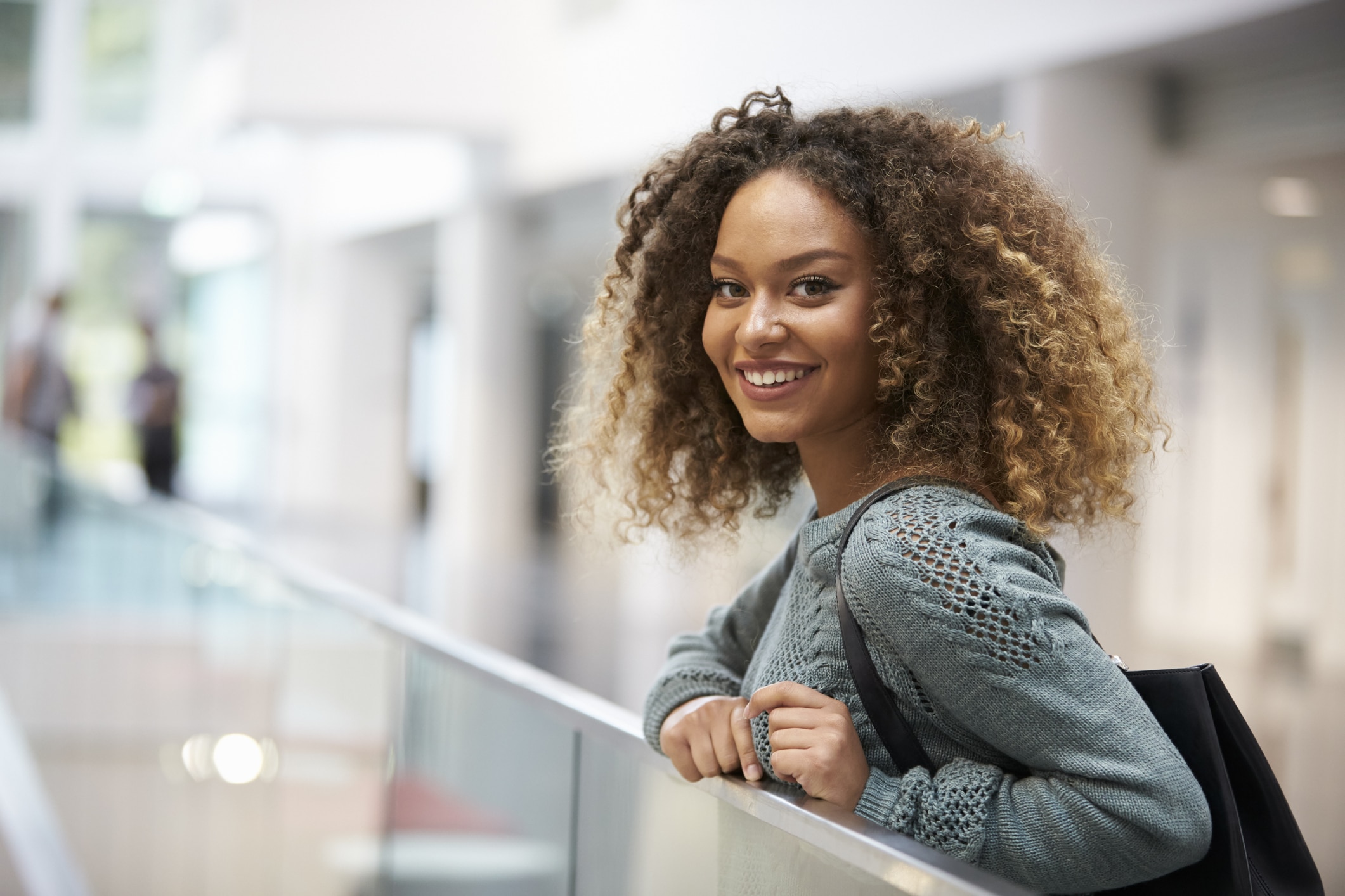  Mulher negra sorridente, com cabelos cacheados, bolsa preta, apoiada em uma grade e olhando para trás