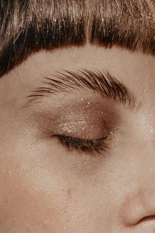 A imagem mostra um rosto feminino, focada em apenas um lado do rosto. A mullher usa uma sombra com brilho nos olhos e a técnica soap brows nas sobrancelhas  
