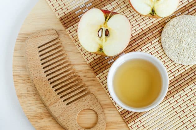 A imagem mostra uma maça cortada ao meio, ao lado de um recipiente com vinagre de maçã e um pente de madeira