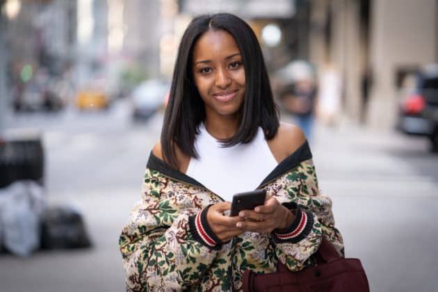 Mulher negra de cabelo alisado, chanel. Ela usa uma blusa branca e uma jaqueta estampada por cima. Carrega uma bolsa marrom e um dos braços e segura um celular com as mãos