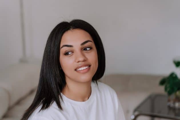 Mulher negra de cabelo alisado, longo, partido ao meio. Ela está vestindo uma camiseta branca, sentada em um sofá, sorrindo