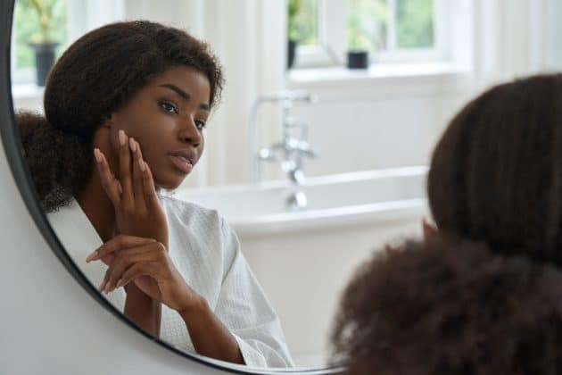 Mulher de pele negra se olhando no espelho enquanto passa a mão pelo rosto