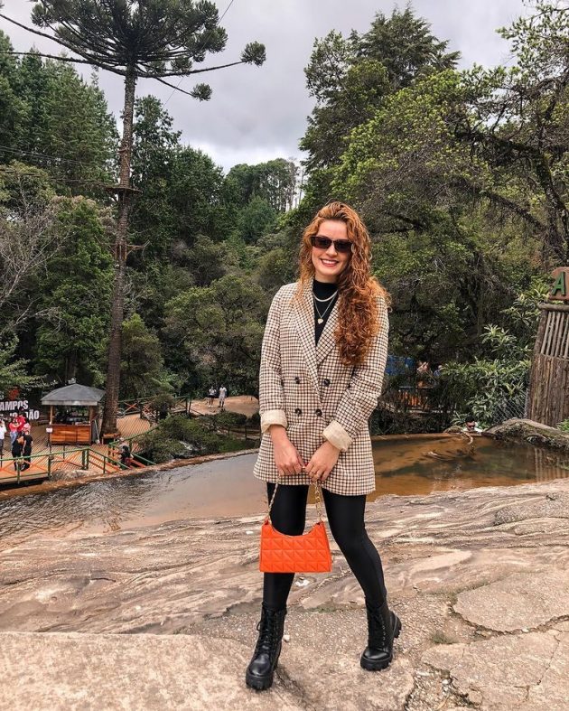 Moça com calça preta, bolsa laranja na mão e sobretudo bege.