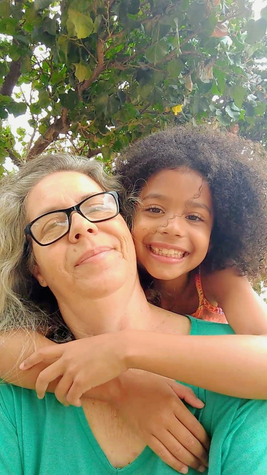  Mãe de óculos abraçada com criança sorridente, ao ar livre com árvores ao fundo