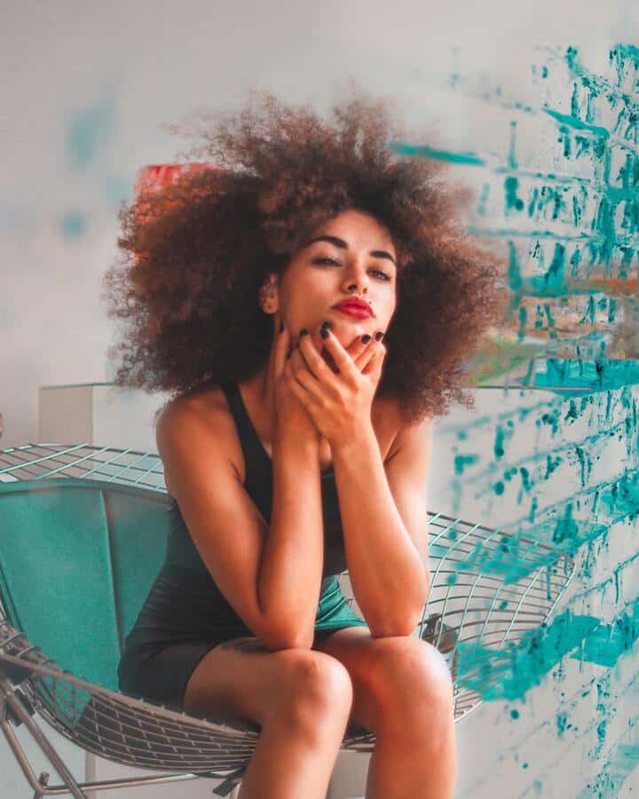  Mulher negra de cabelo crespo castanho, super volumoso, com aspecto saudável e brilhoso (benefício do ácido hialurônico no cabelo). Ela está sentada em uma cadeira verde, usa um vestido justo cinza e batom vermelho