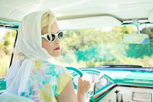 Mulher branca, de cabeo loiro (envolvido em um lenço branco), com óculos escuros e roupas na estética old money, dirigindo um carro antigo