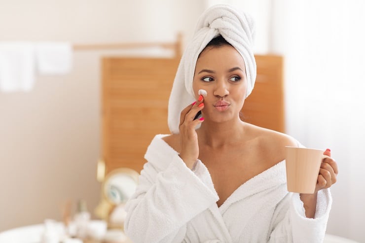  Mulher negra com toalha branca na cabeça, vestindo um roupão branco caído nos ombros, com uma xícara de café em uma mão e passando um creme de tratamento de pele no rosto com a outra mão