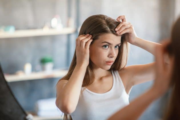 Mulher branca de cabelo liso, castanho claro, em frente ao espelho observando o couro cabeludo ressecado