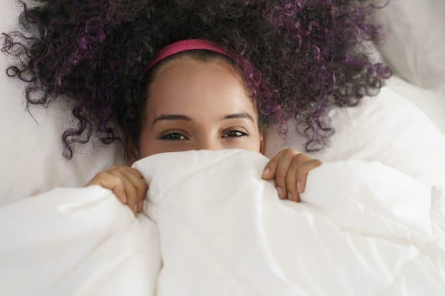 Mulher de cabelo cacheado, castanho escuro com mechas roxas. Ela está deitada na cama, com o edredom cobrindo o rosto na altura dos olhos