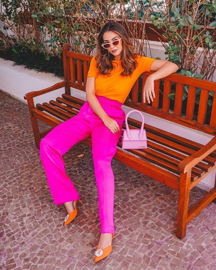  Mulher jovem, com look no estilo da tendência dopamine dressing. Ela veste uma t-shirt laranja e uma calça de alfaiataria rosa pink. Nos pés ela aposta em um sapato scarpin laranja e para compor o look, uma bolsa e um óculos escuro, ambos na cor rosa bebê