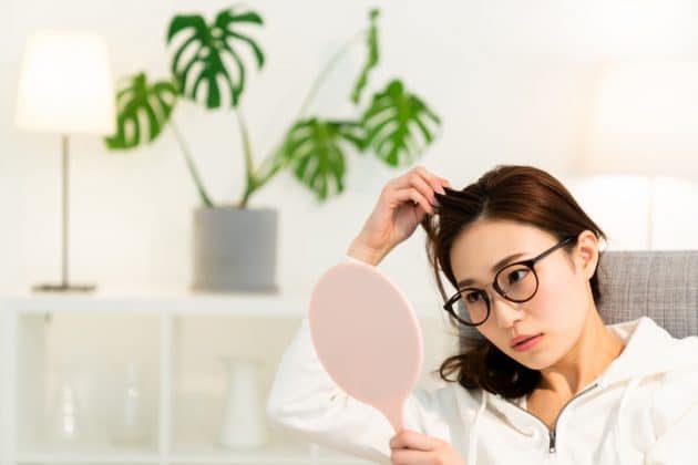 Mulher branca de cabelo liso e castanho, coçando o couro cabeludo em frente ao espelho