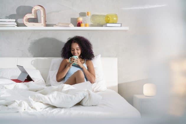 Mulher jovem, negra, de cabelo crespo, corte acima do ombro. Ela está sentada na cama, tomando uma xícara de café