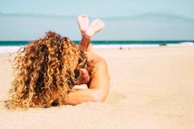 Mulher de cabelo cacheado loiro, longo. Ela está na praia, deitada de bruço na areia