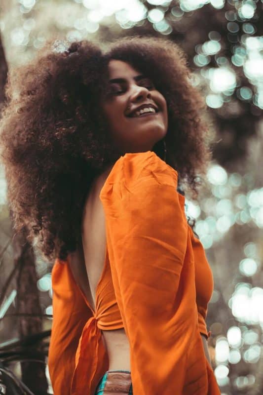Mulher de cabelo crespo, longo e volumoso. Ela está vestindo um cropped de manga longa, laranja