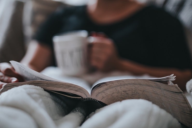  A imagem mostra uma pessoa sentada na cama, lendo um livro e tomando uma xícara de café