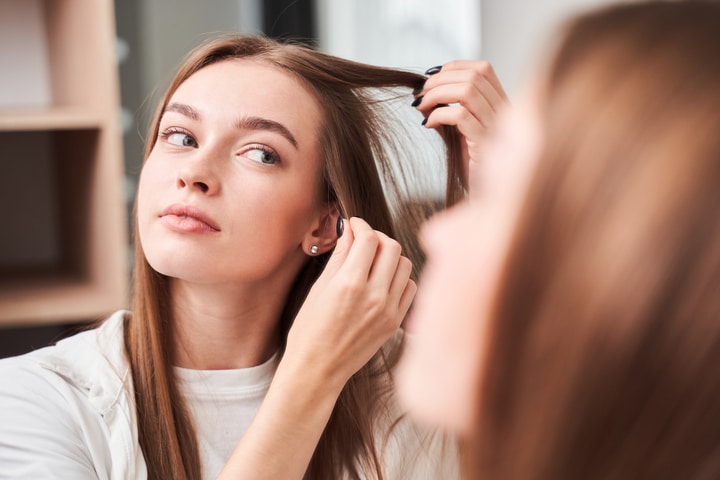 Mulher de cabelo loiro, liso e longo, observando uma mecha no espelho