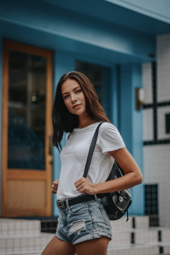 Mulher com look de verão: camiseta branca básica, short jeans com cinto preto e bolsa preta, no estilo mochilinha