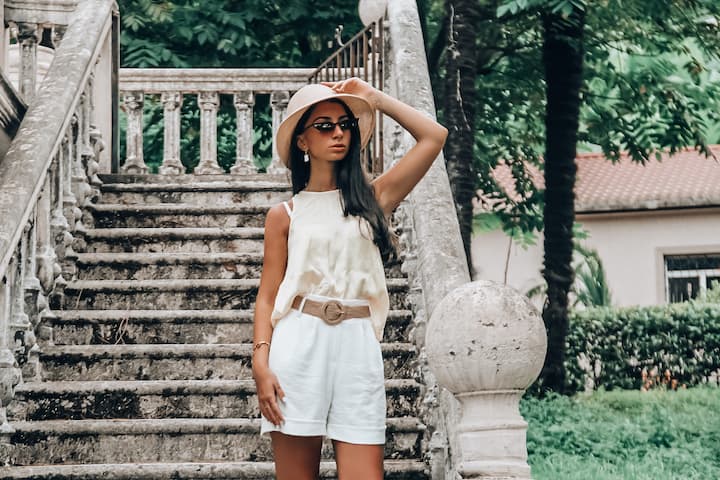 Mulher com blusa bege, short de alfaiataria branco, com um cinto marrom, óculos escuro e chapéu branco