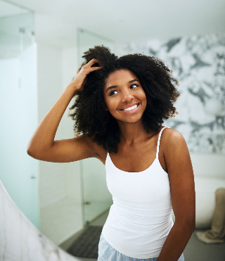  Jovem com cabelo cacheado em um ambiente claro, sorriso radiante, interagindo com seu cabelo na frente do espelho. Perfeita para dicas de cuidados com cabelo.