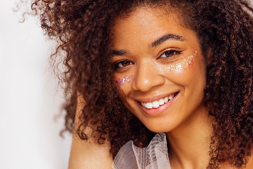 Uma jovem mulher sorridente com cabelos cacheados e maquiagem brilhante, destacando-se com glitter no rosto. Ela exibe uma expressão alegre e confiante.