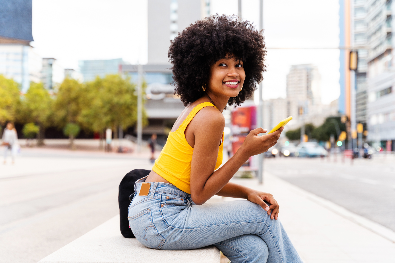 Jovem sorridente com cabelo afro usando uma blusa amarela e jeans sentada em um banco em ambiente urbano, segurando um celular.