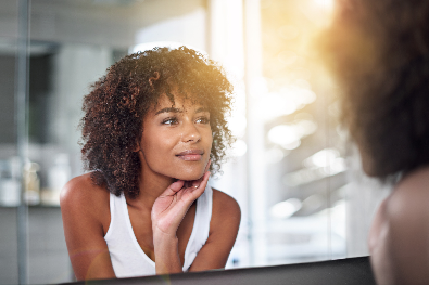 Uma mulher com cabelo crespo observa seu reflexo no espelho, refletindo autoconfiança e beleza natural em um ambiente iluminado.
